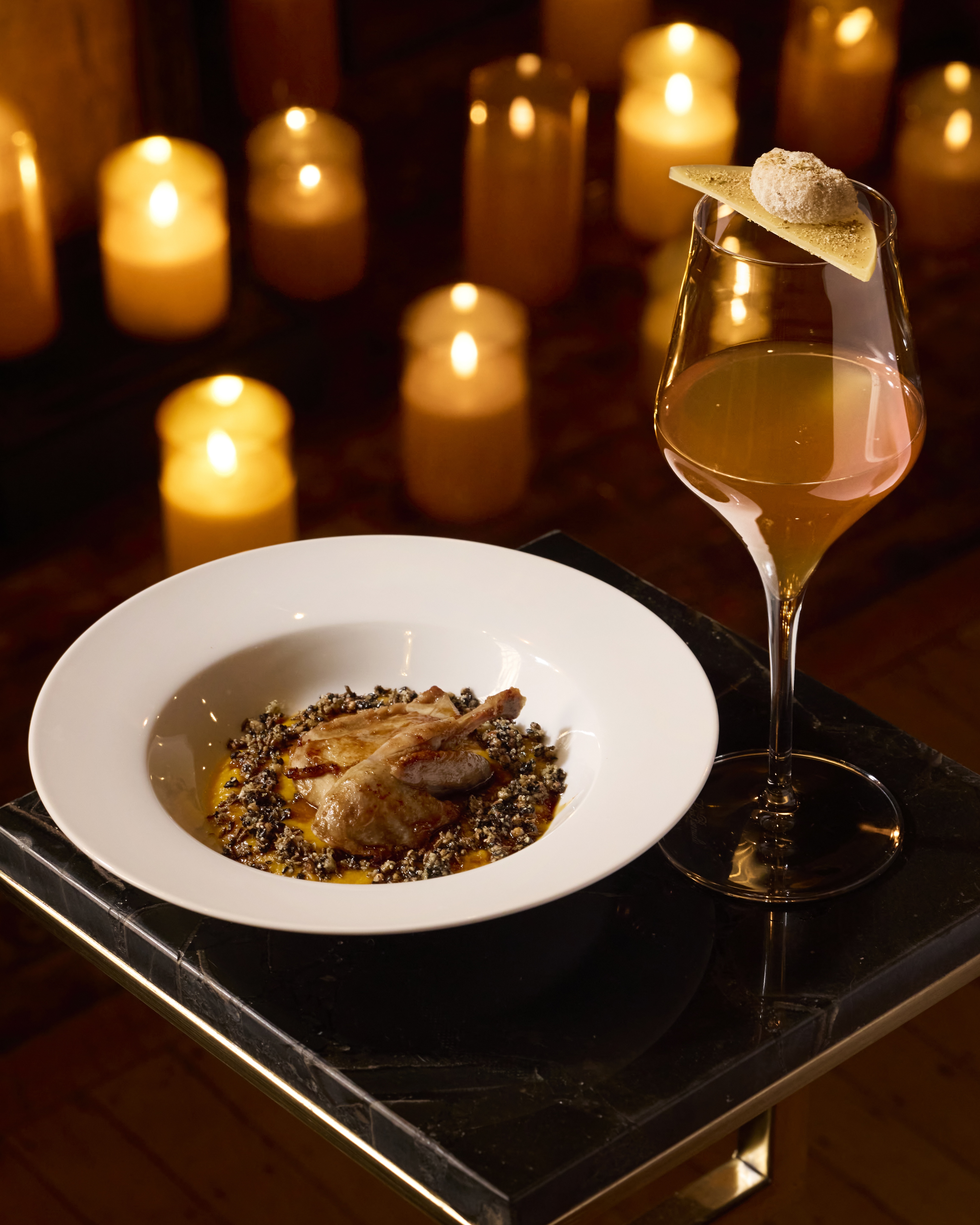 An elegant cocktail in a stemmed glass beside a plated dish, bathed in warm candlelight